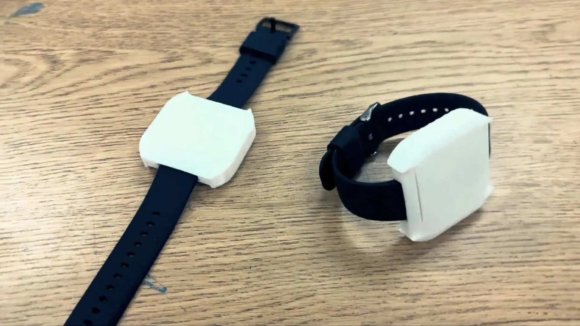 Two white 3D-printed wristband prototypes on black watch straps, lying on a wooden table