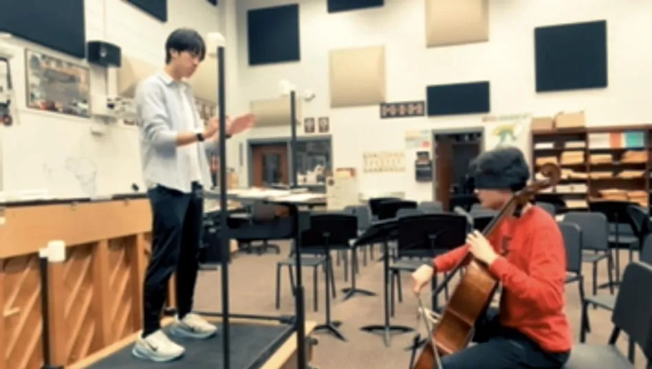 A conductor on a podium inside a frame of four posts while a blindfolded cellist plays
